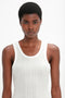 A person with short curly hair is wearing a white breathable lightweight ribbed knit sleeveless top, reminiscent of Victoria Beckham's Fine Knit Vertical Stripe Tank In White, and is looking straight ahead with a neutral expression.