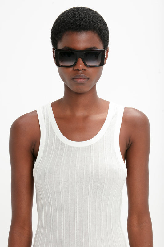 A person wearing a white sleeveless top and Large Black Oversized Frame Sunglasses In Black with Victoria Beckham’s signature B frame buckle stands against a plain white background.