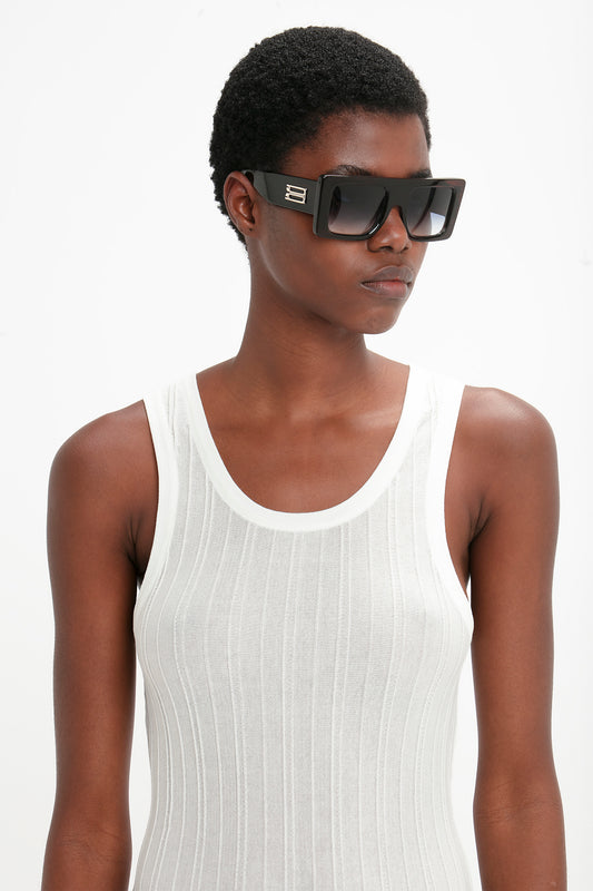 A person with short hair wearing bold, Oversized Frame Sunglasses In Black by Victoria Beckham and a white ribbed tank top stands against a plain white background.