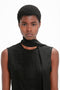 A person with short curly hair wears a Sleeveless Tie Neck Top In Black by Victoria Beckham and a matching scarf made of crepe de chine silk, looking straight ahead against a plain white background.