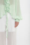 Close-up of a person wearing a Victoria Beckham Romantic Blouse In Jade, featuring ruffled details and matching translucent white pants. The jade hue subtly accentuates the diaphanous blouson sleeves while the left arm and hand are visible, showcasing a buttoned cuff.