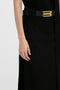 Close-up of a person wearing a sophisticated black outfit with a wide black belt featuring a large gold buckle. The Tailored Floor-Length Skirt In Black by Victoria Beckham adds an elegant touch, while the person's left hand hangs by their side. The background is plain white.