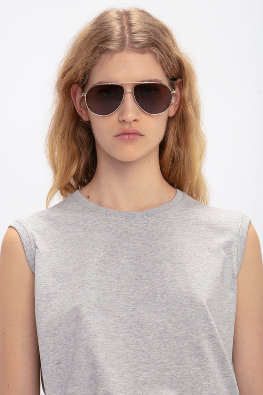 A person with long blonde hair wearing Victoria Beckham V Metal Pilot Sunglasses In Gold-Khaki with adjustable nose pads and a sleeveless gray top is looking straight ahead with a neutral expression.