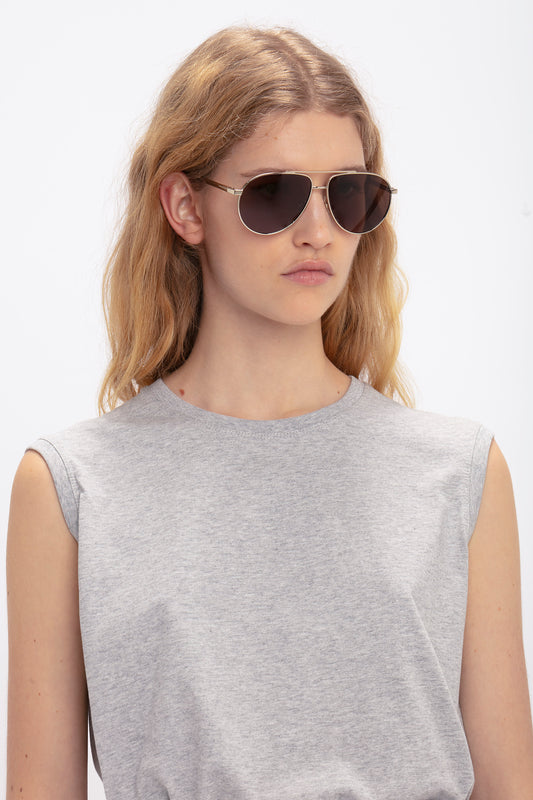 A person with long, wavy hair is wearing Victoria Beckham V Metal Pilot Sunglasses In Gold-Khaki featuring adjustable nose pads, complemented by a sleeveless gray top, set against a plain, white background.