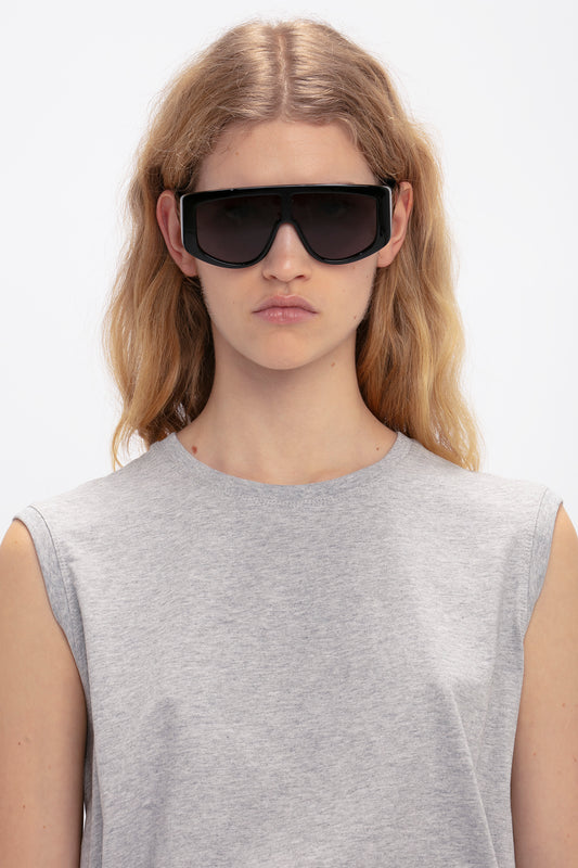 A person with long blonde hair is wearing Victoria Beckham Acetate Visor Sunglasses In Black featuring oversized frames and black tonal lenses, along with a sleeveless gray top, looking directly at the camera on a plain white background.