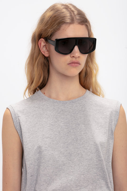 A person with long blonde hair wearing Victoria Beckham Acetate Visor Sunglasses In Black featuring oversized frames and black tonal lenses stands against a plain background, complemented by a light gray sleeveless top.