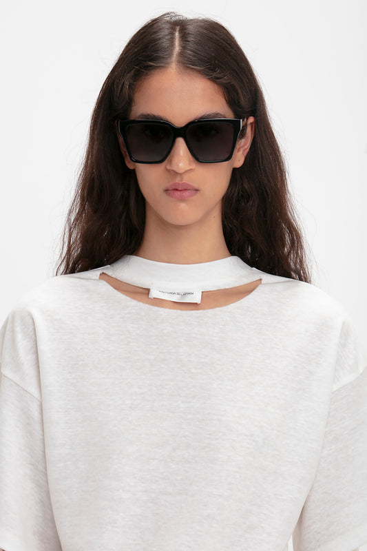 A woman with long dark hair, wearing Victoria Beckham Soft Square Frame Sunglasses In Black and a white Bonded Jersey Tee featuring cut-out detailing on the neckline, stands against a plain white background.