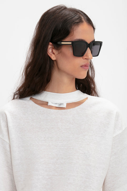 A person with long dark hair is wearing large Victoria Beckham Soft Square Frame Sunglasses In Black and a light-colored, cutout sweatshirt. The background is plain white.