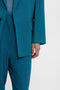 Close-up of a person wearing a Victoria Beckham brand teal suit with matching blazer and trousers. The right hand hangs by the side, revealing a light blue top under the Victoria Beckham Peak Lapel Jacket In Petroleum. The oversized silhouette adds a contemporary touch to the ensemble.