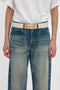 Person wearing a white t-shirt and blue jeans with a beige Victoria Beckham Jumbo Frame Belt In Latte Leather featuring gold hardware, standing with arms by their sides against a plain background.