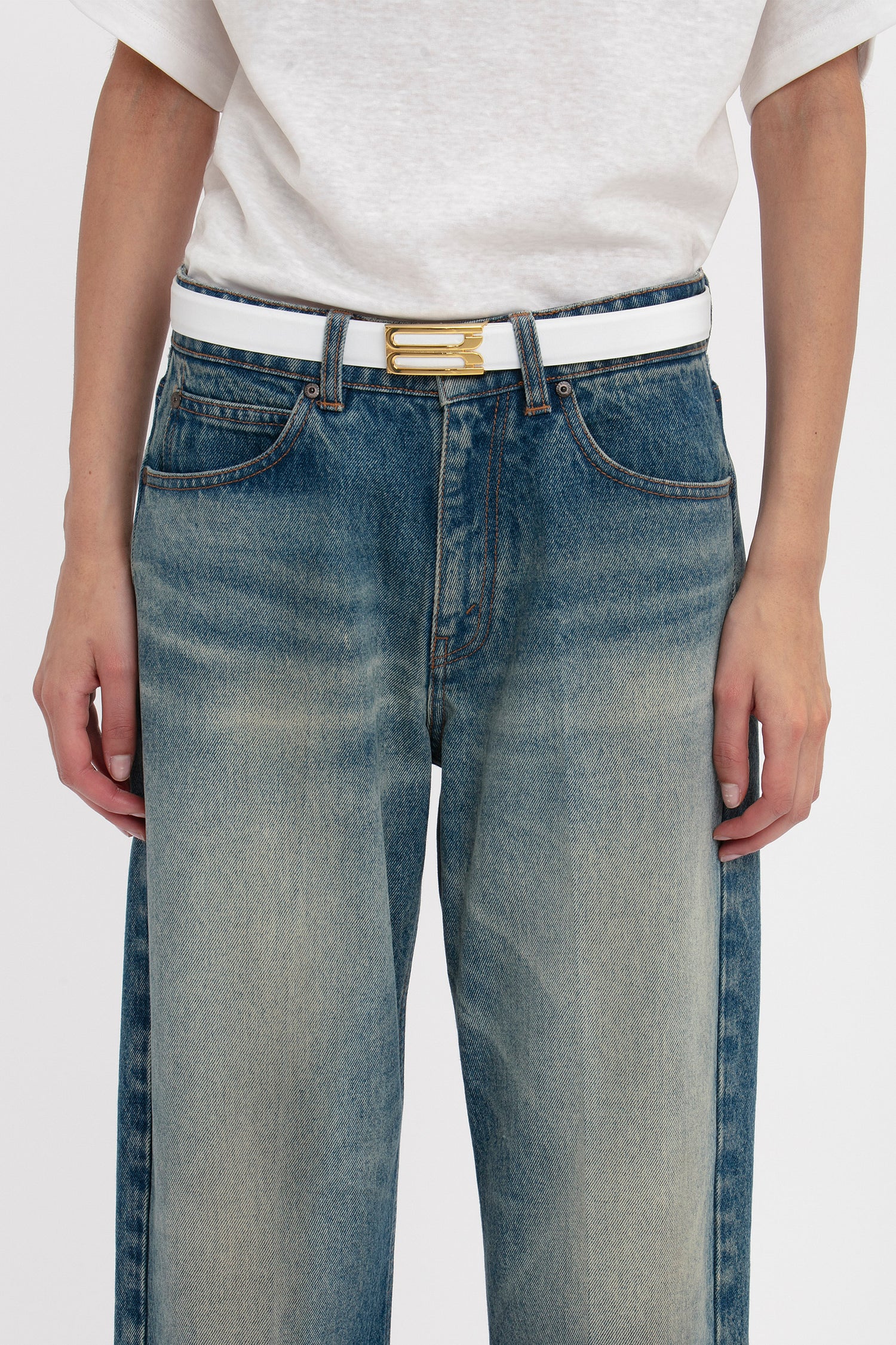 A person wearing a white t-shirt, blue jeans, and a Victoria Beckham Frame Belt In White Leather featuring gold hardware. The image is cropped to show only the torso and legs.