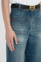 Close-up of a person wearing vintage denim Victoria Beckham Alina High Waisted Jean In Indigrey Wash with front pockets, a black belt with a gold buckle, and a white shirt. The jeans feature a small red embroidered logo on the left pocket, adding a touch of seventies-style flair.