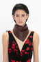 A person with short dark hair is wearing a black sleeveless dress with red floral patterns and a Leather Scarf In Bordeaux by Victoria Beckham. They are facing forward against a plain white background, showcasing an elegant blend of feminine separates and masculine tailoring.