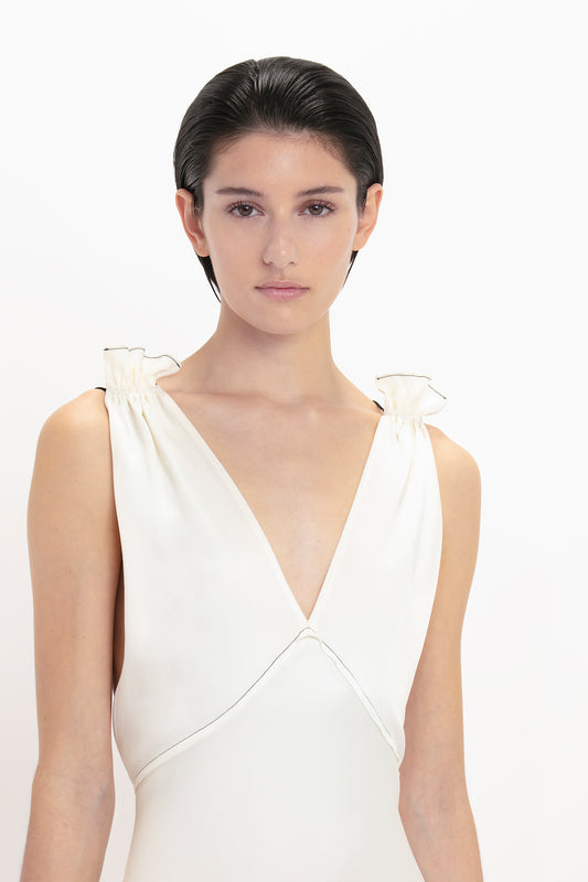 A person with short, dark hair is shown against a plain background, wearing a white, sleeveless Gathered Shoulder Floor-Length Cami Gown In Ivory made of crepe back satin with intricate gathered shoulder details from Victoria Beckham.