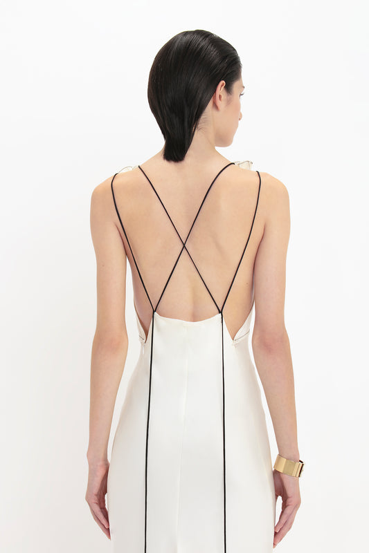 A person with dark hair, wearing a backless Gathered Shoulder Floor-Length Cami Gown In Ivory by Victoria Beckham made of crepe back satin with black cross straps, stands facing away from the camera against a plain white background.