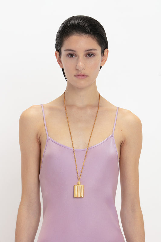 A person with short dark hair wearing a Victoria Beckham Low Back Cami Floor-Length Dress In Rosa and a long gold necklace with a rectangular pendant, standing against a plain white background.