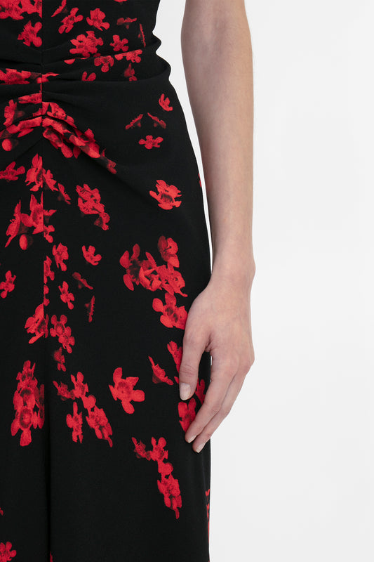Close-up of a person’s right arm and hand resting at their side, wearing a Gathered Waist Midi Dress In Sci-Fi Black Floral with a red print. The dress has a gathered detail at the waist, showcasing its fit-and-flare silhouette from Victoria Beckham.
