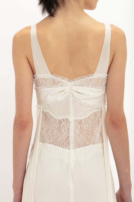 A woman is wearing the Victoria Beckham Ruffle Detail Midi Dress in Ivory with thin straps, viewed from the back, showing intricate lace panels and a fabric bow design on the upper back.