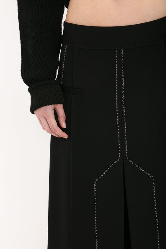 Close-up of a person wearing a black long-sleeved top and a Victoria Beckham Deconstructed Floor-Length Skirt In Black with white stitching details. The person's left hand rests on their hip, accentuating the elongating silhouette.