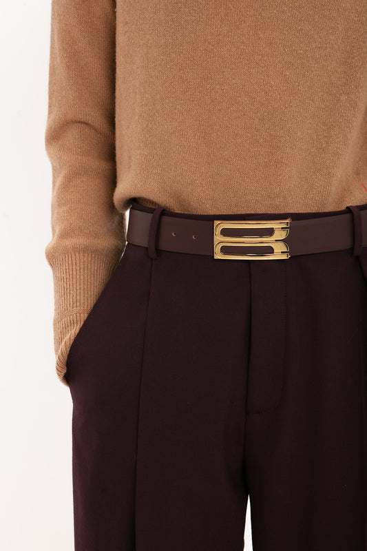 Person wearing a brown sweater tucked into Victoria Beckham Asymmetric Chino Trouser In Deep Mahogany with hands in pockets, featuring a burgundy belt with a large gold buckle.