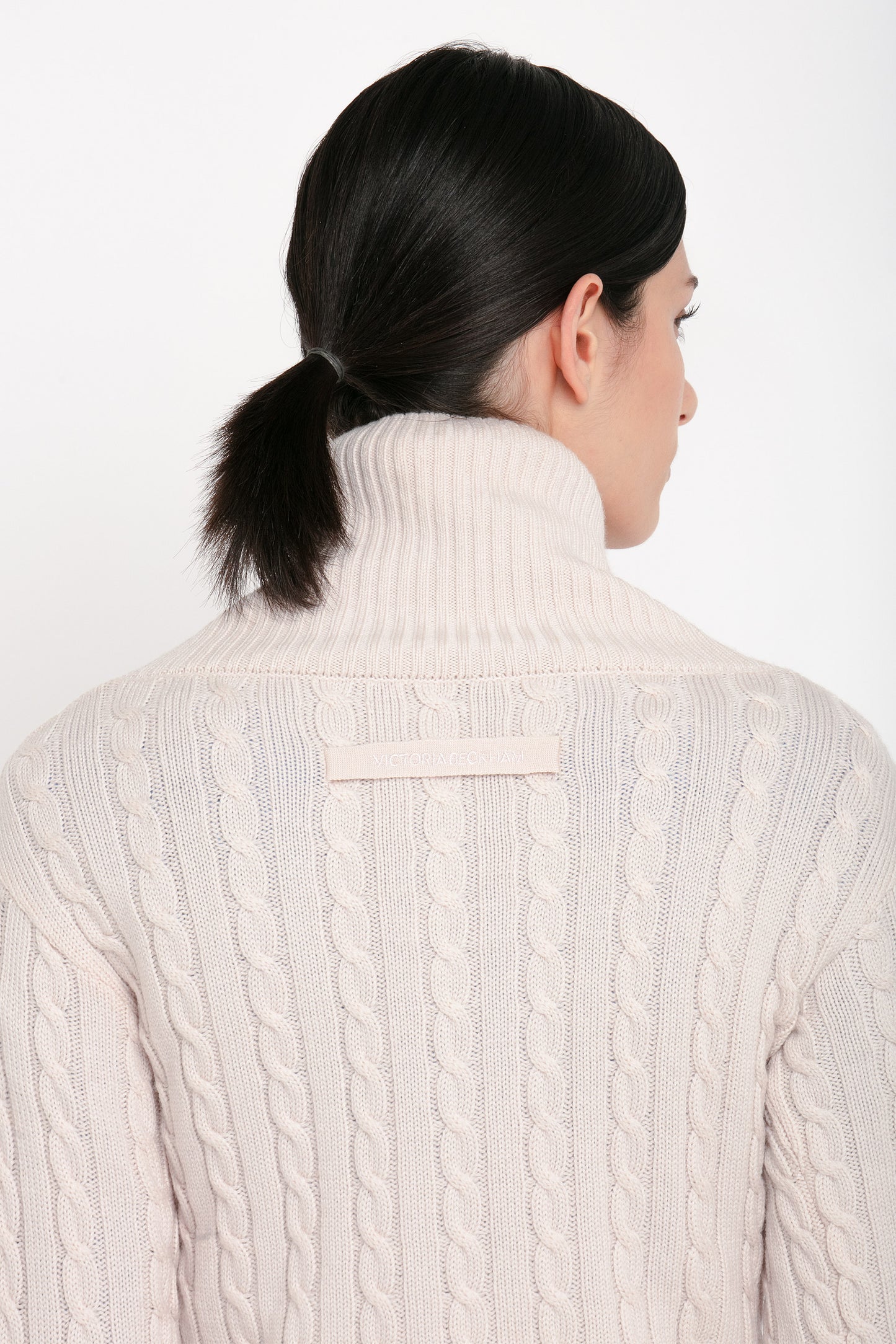 A person with dark hair tied in a low ponytail is shown from the back, wearing a high-neck, cream-colored Wrap Detail Jumper In Bone by Victoria Beckham made from luxurious merino wool.