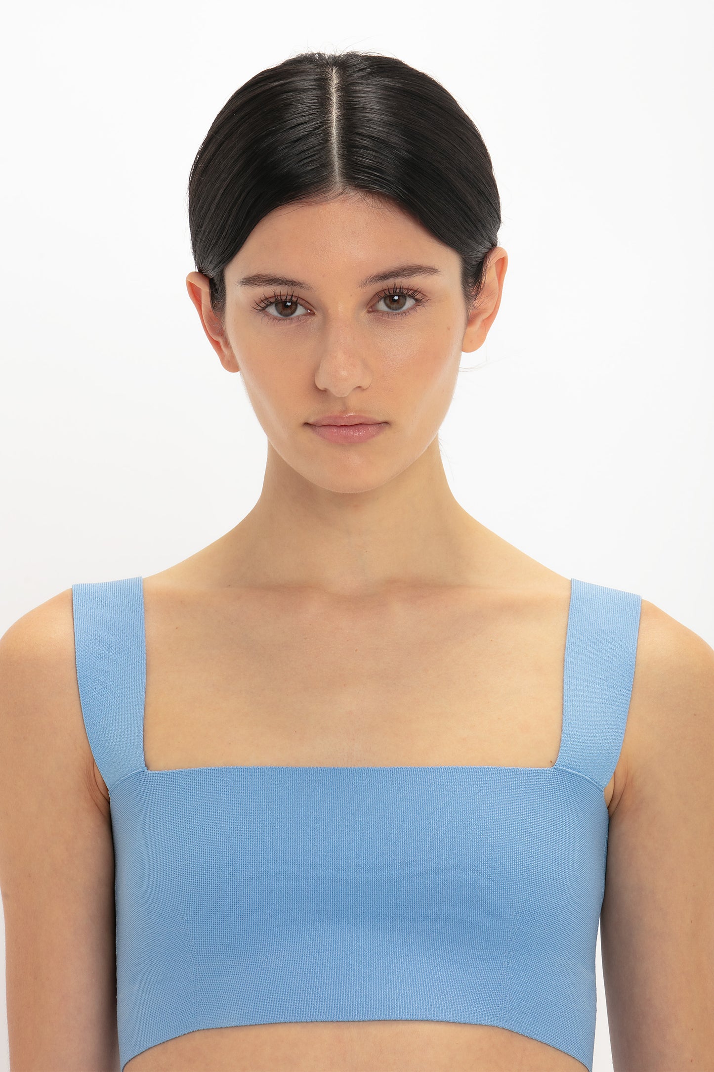 A person with dark hair in a center part is wearing a light blue Strap Bandeau Top In Marina by Victoria Beckham, facing forward against a white background.