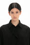 A person with dark hair, wearing a Victoria Beckham Asymmetric Ruffle Blouse In Black that exudes contemporary timeless fashion, looks into the camera against a plain white background.