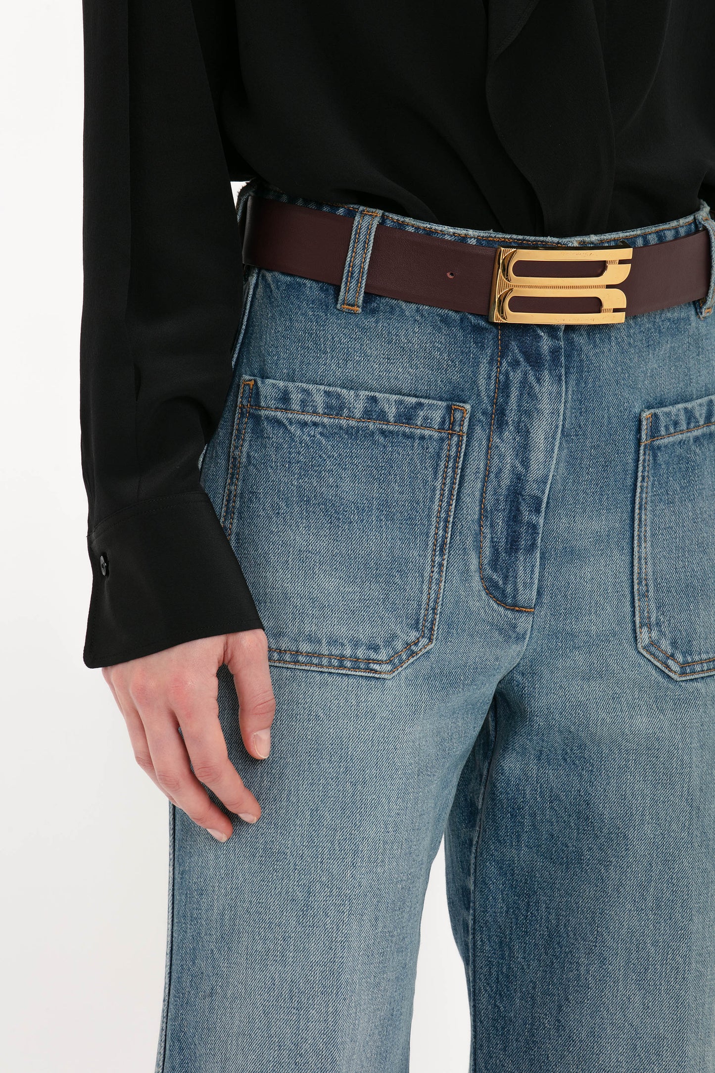 Close-up of a person wearing a black shirt tucked into high-waisted Victoria Beckham Alina High Waisted Jean In Worn Blue Wash with large front pockets, accessorized with a brown belt featuring a gold buckle.