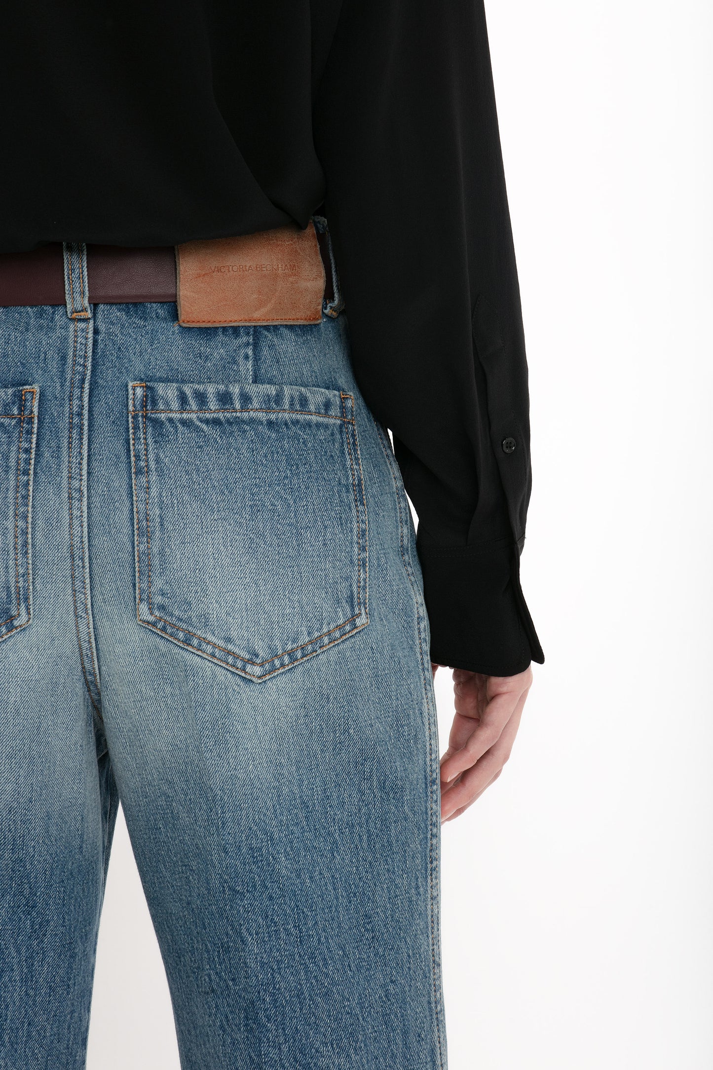 A person wearing distressed Alina High Waisted Jean In Worn Blue Wash by Victoria Beckham with a brown leather belt and a black shirt, photographed from the back.