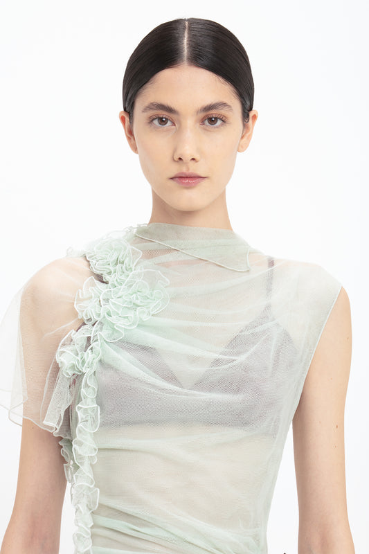 A person with dark hair tied back wears a light green, semi-sheer, one-shoulder top adorned with ruffled fabric accents. This limited edition piece is reminiscent of the Victoria Beckham Gathered Tulle Detail Floor-Length Dress In Jade with its elegant sea foam green tulle details.