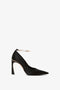 A single black high-heeled shoe with a pointed toe, featuring a slim 100mm heel and an elegant ankle strap adorned with a delicate metal ankle bracelet. The Victoria Beckham Pointy Toe Pump in Black Satin classic pump design exudes sophistication and style.