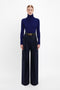 A person stands against a plain white background, wearing a dark blue turtleneck sweater and Wide Leg Trouser In Midnight by Victoria Beckham, secured with a black belt.