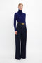 A person stands against a plain background wearing a blue turtleneck, Victoria Beckham Wide Leg Trouser In Midnight, and a black belt.