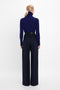 A person facing away from the camera wears a dark blue turtleneck sweater and high-waisted navy **Wide Leg Trouser In Midnight by Victoria Beckham**, with their hair pulled back.