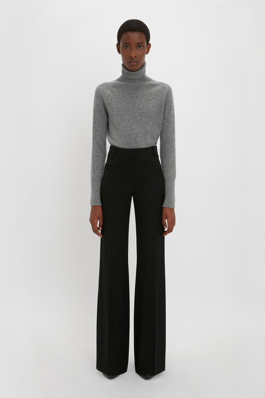 Person standing against a plain white background, wearing a Polo Neck Jumper In Grey Melange and high-waisted black pants with the Victoria Beckham monogram. They have a neutral expression and arms relaxed at their sides, showcasing versatile styling options.