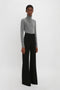 Person standing against a plain white background wearing a gray turtleneck sweater and Alina Tailored Trouser In Black by Victoria Beckham that evoke 70s style references.