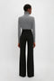 A person with short hair is seen from behind, wearing a gray turtleneck sweater and Alina Tailored Trouser In Black by Victoria Beckham that evoke subtle 70s style references.