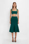 A woman stands in a fitted green crop top and matching VB Body Flared Skirt In Lurex Green by Victoria Beckham, featuring a flattering flared silhouette, paired with black high-heeled sandals, against a plain white background.