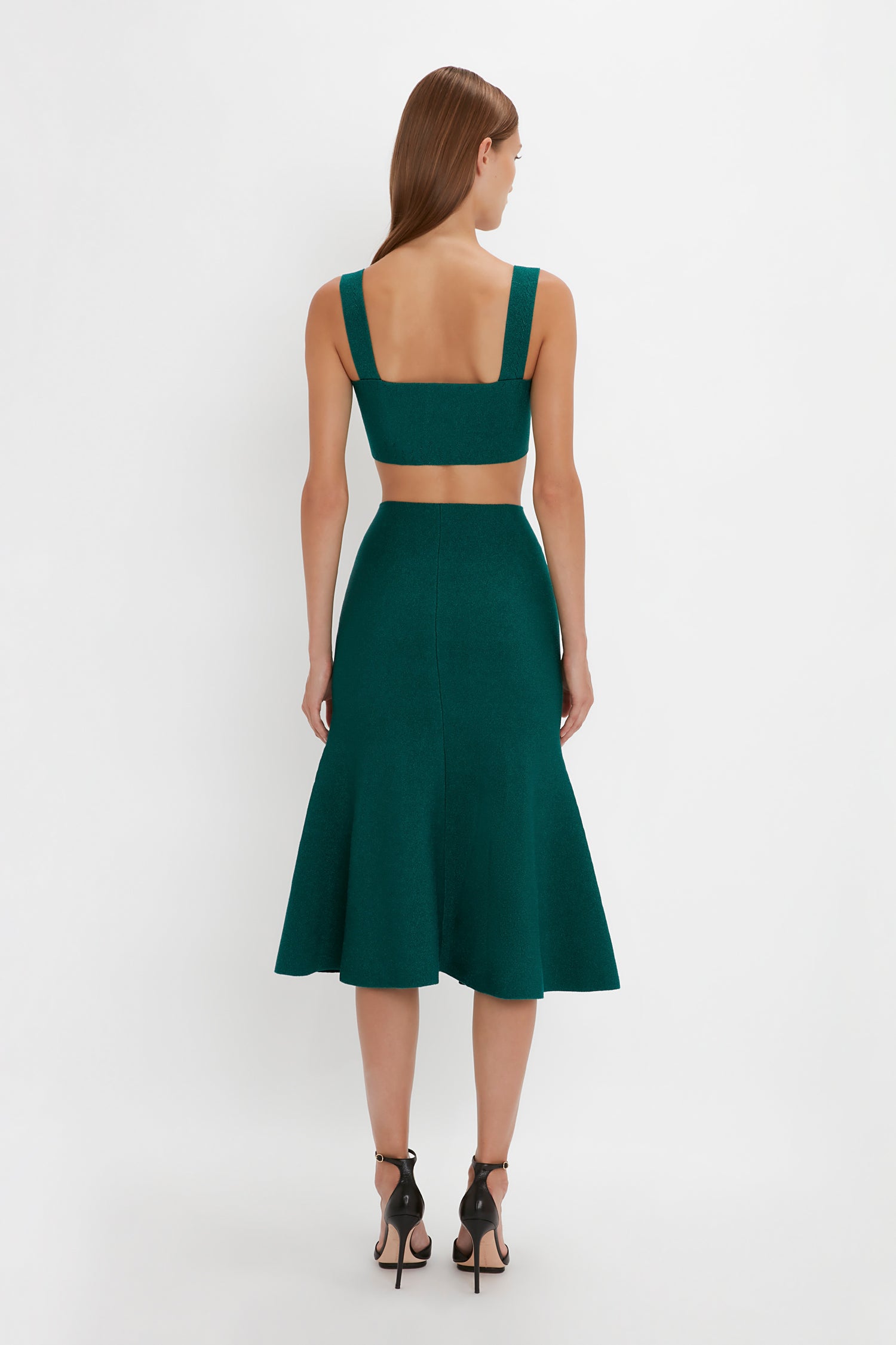 A woman wearing a VB Body Flared Skirt In Lurex Green by Victoria Beckham, viewed from the back. She has long brown hair and is standing against a plain white background.