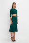 A woman stands in a Victoria Beckham VB Body Cropped Cardi In Lurex Green and matching high-waisted skirt, both accentuating her form-fitting silhouette and feminine sensibility, paired with black high heels, against a plain white background.