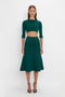 Person wearing a dark green, buttoned VB Body Cropped Cardi In Lurex Green by Victoria Beckham and a matching flared skirt, the form-fitting silhouette exuding a feminine sensibility against a plain white background.