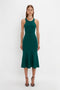 A woman stands against a white background, wearing a Victoria Beckham VB Body Sleeveless Dress In Lurex Green. The flattering flared silhouette enhances her figure, while her black high-heeled sandals add the perfect touch to this new-season wardrobe essential.