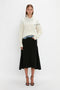 A person stands against a plain background wearing the Collar Detail Jumper In Natural by Victoria Beckham with casual sophistication, complemented by a black skirt and black heels.