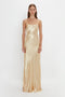 A person wearing a 1990s-inspired, long, silky Exclusive Floor-Length Cami Dress In Gold with thin straps by Victoria Beckham, crafted from crepe back satin, standing against a plain white background.