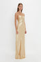 A person stands on a white background, wearing the Exclusive Floor-Length Cami Dress In Gold by Victoria Beckham, crafted from luxurious crepe back satin with delicate spaghetti straps, exuding a chic 1990s-inspired elegance.