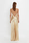 A woman stands with her back to the camera, wearing the Victoria Beckham Exclusive Floor-Length Cami Dress In Gold. The gold shade crepe back satin fabric catches the light beautifully as her straight, brown hair falls just above her shoulders.