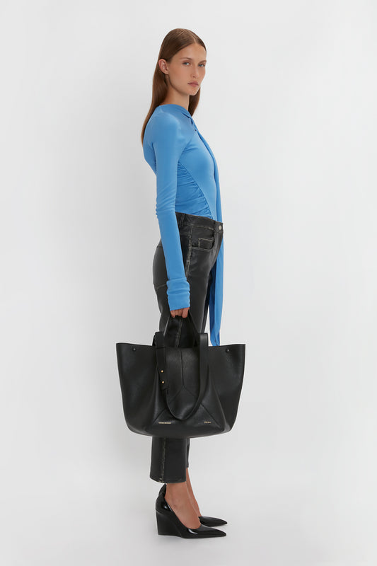 A person in a blue top and black leather pants stands holding a luxurious Victoria Beckham W11 Medium Tote In Black Leather, wearing black heeled shoes.