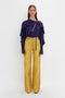 A person stands against a white background, wearing a purple Tie Detail Ruffle Blouse In Ultraviolet by Victoria Beckham with a V neckline and yellow wide-leg pants featuring a striped pattern.