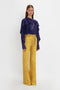 A person stands against a plain white background, wearing a Victoria Beckham Tie Detail Ruffle Blouse In Ultraviolet with a V neckline and high-waisted yellow pants featuring a subtle striped pattern.