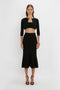 A person stands against a white background wearing a Victoria Beckham VB Body Cropped Cardigan In Black over a black bandeau top and a black mermaid skirt, showcasing a form-fitting silhouette enhanced by black high heels.